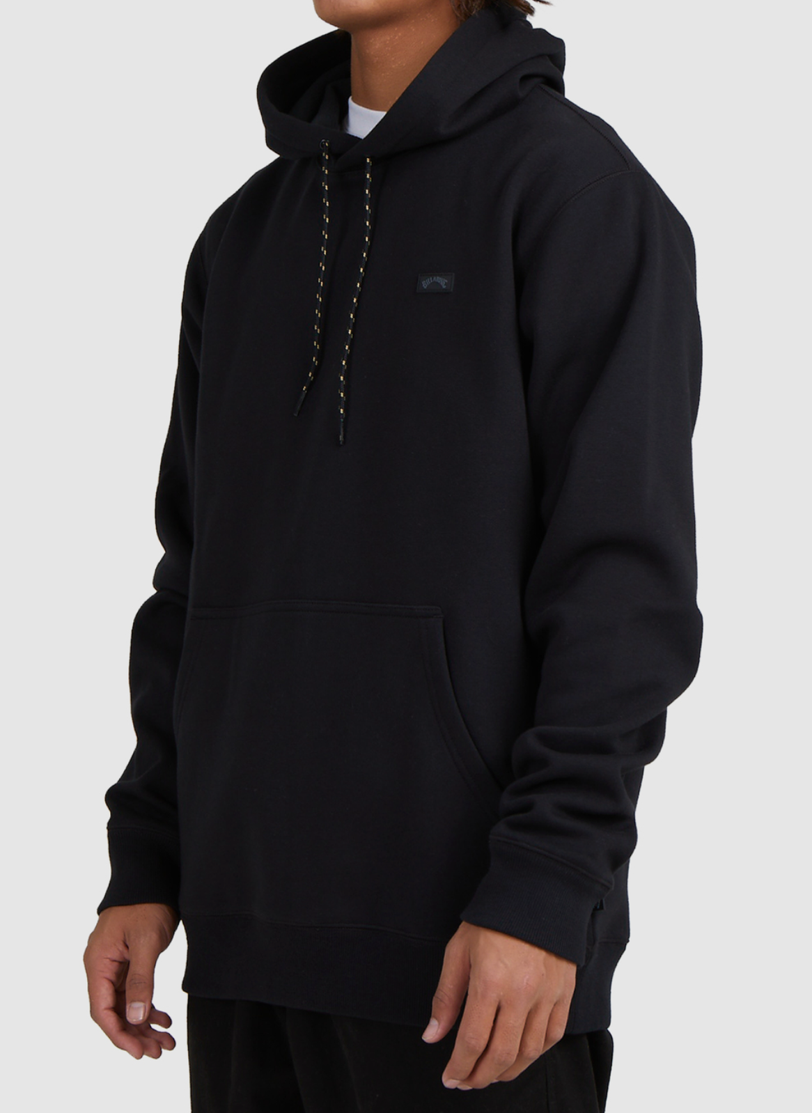 A person in a black Billabong Shoreline Pullover with a front pocket and black drawstrings stands against a plain light background. The heavyweight casual hoodie is shown from the upper body only, with the face out of view.