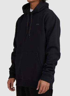 A person in a black Billabong Shoreline Pullover with a front pocket and black drawstrings stands against a plain light background. The heavyweight casual hoodie is shown from the upper body only, with the face out of view.