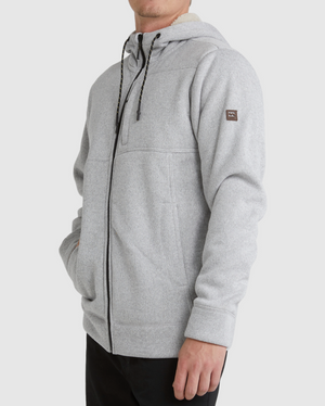 Someone wearing the Billabong Boundary Sherpa Zip in light gray, featuring black drawstrings, a small logo patch on the upper left sleeve, and warm sherpa lining, stands against a plain light background.