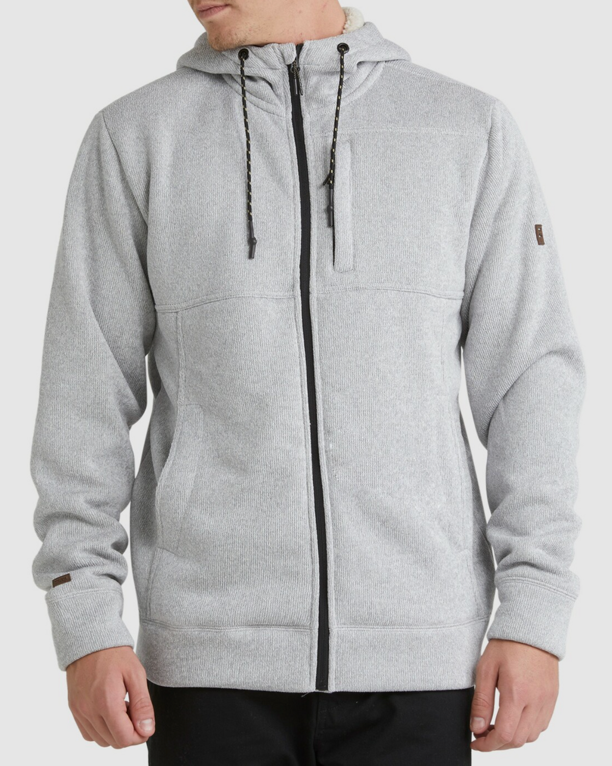 A person wears the Billabong Boundary Sherpa Zip in light gray with black drawstrings, front pockets, and black pants, standing against a plain light background. The hoodie is lined with cozy sherpa for extra warmth.