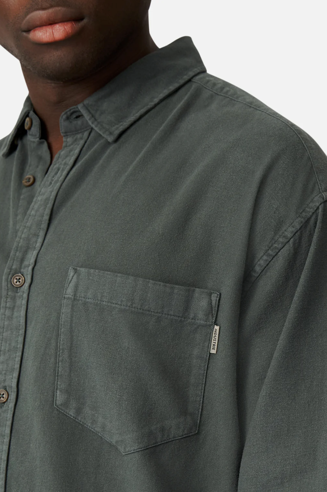 A close-up showcases a person wearing the Industrie "The Taylor Shirt" in dark green, made from 100% cotton. It features brown buttons, a chest pocket with the Industrie logo on a tag at the seam, set against a plain white background.