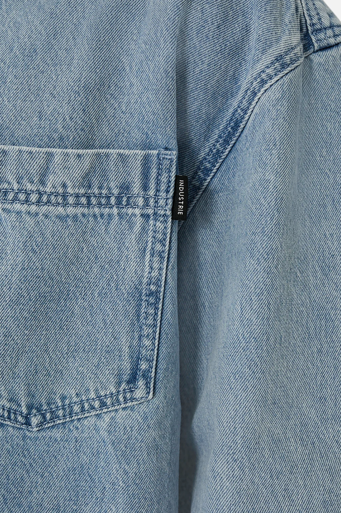 Close-up of Industrie The Lafayette Shirt in light blue denim, relaxed fit, with a visible back pocket and a small black tag stitched on the seam next to it.