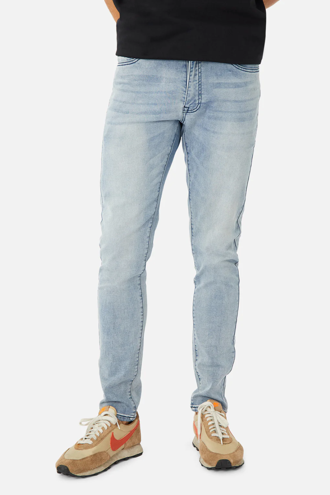 A person is wearing light blue Industrie The Denim Drifter NC Pant jeans, a black shirt, and tan and cream sneakers with orange swooshes. The focus is on the lower half of the body, including legs and feet, against a plain white background.