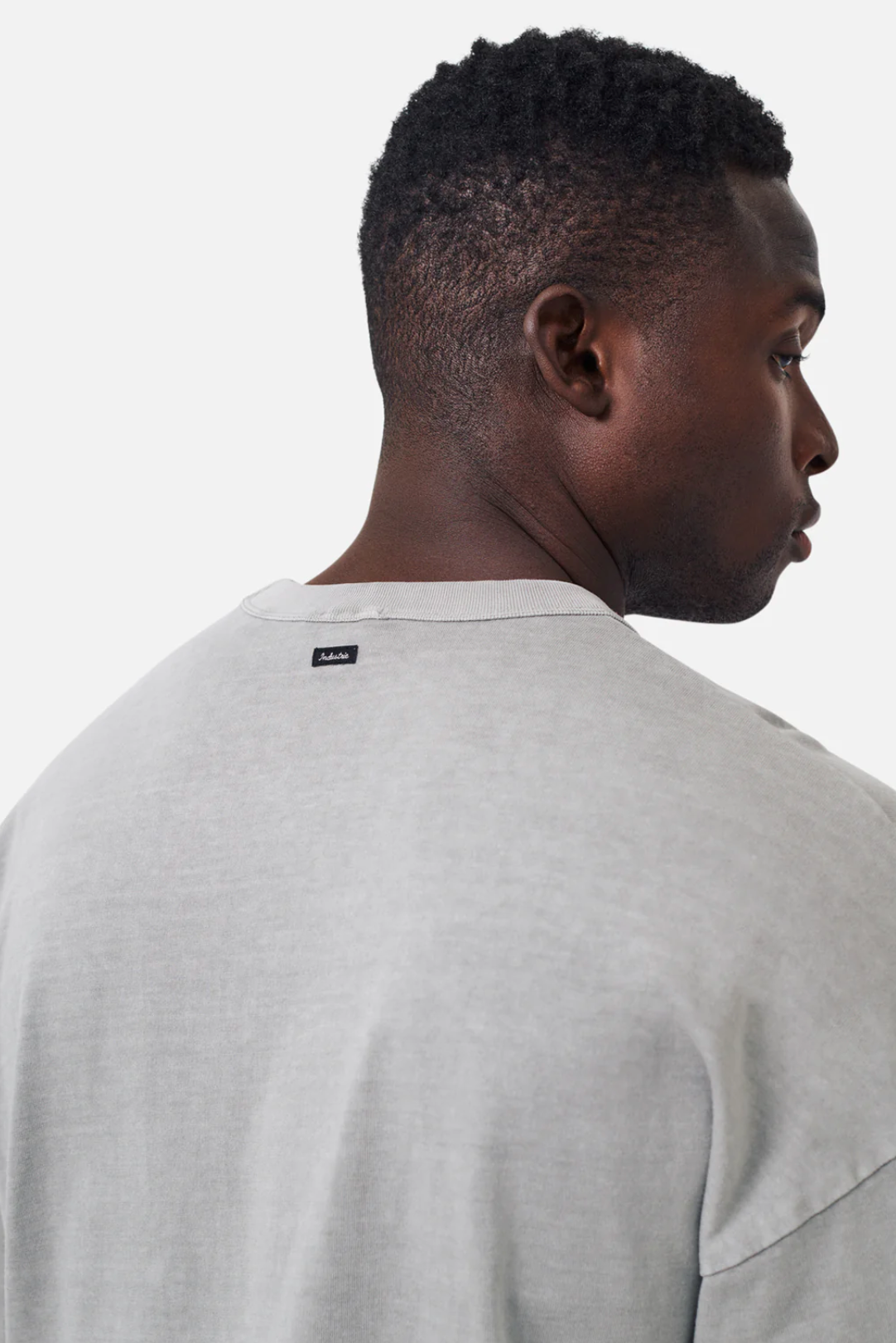 A person with short curly hair is seen from the back wearing an oversized fit, light gray Industrie The Del Sur Long Sleeve Tee, made from 100% cotton. The casual T-shirt has a small black tag on the upper back against a plain white background.