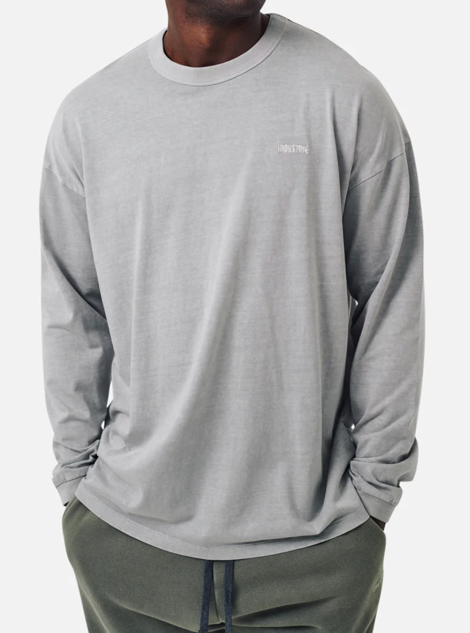 A person wears the Industrie The Del Sur Long Sleeve Tee in light gray, 100% cotton, featuring "Industrie" on the chest. They pair it with oversized dark green pants while posing against a plain white backdrop.