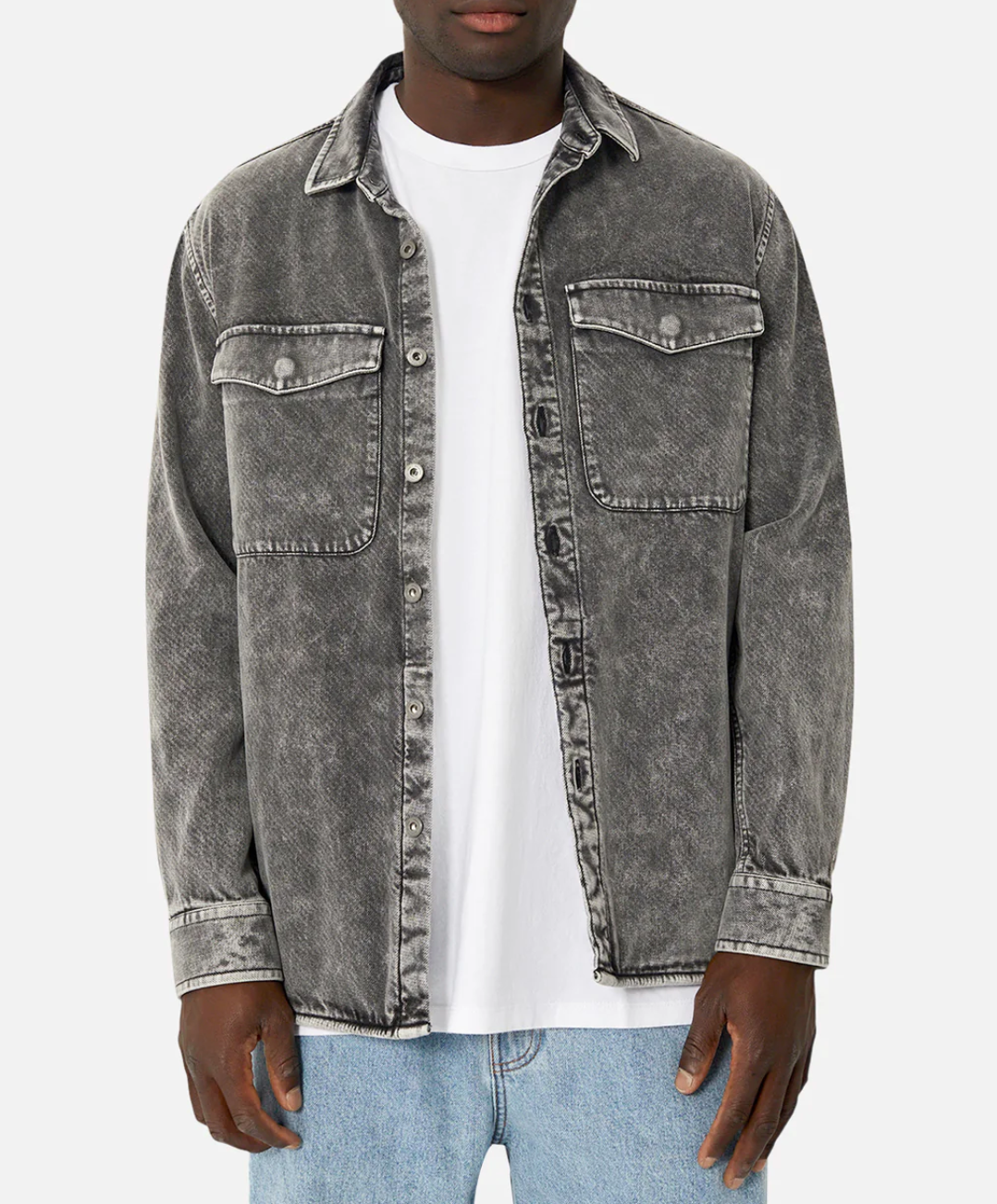 A person is wearing the Industrie The Duster Jacket, a stylish, faded black denim piece made entirely of 100% cotton. It features two chest pockets with flaps and buttons. They have paired it with a white t-shirt and light blue jeans, standing against a plain white background with their hands relaxed by their sides.
