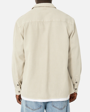 Rear view of a person wearing Industrie The Duster Jacket, a relaxed fit, beige button-up shirt with long sleeves, buttoned cuffs, and a collar. Made from 100% cotton and stylishly paired with light blue jeans against a plain white background.