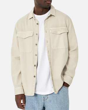 An individual is wearing the Industrie The Duster Jacket, a relaxed fit light beige button-up made from 100% cotton, layered over a white t-shirt and blue jeans against a plain white background. It has two chest pockets with flaps. The person's face remains unseen.