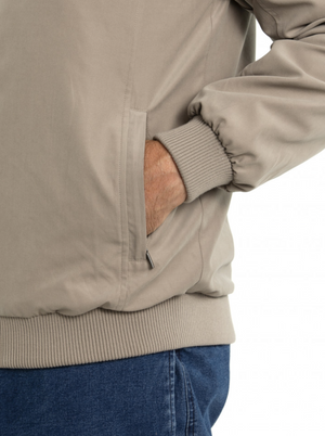 The individual sports the Breakaway Microfibre Bomber Jacket in beige, featuring a ribbed hem and cuffs with zippered pockets. Their hand rests comfortably inside the jacket pocket, paired with blue jeans. The photo frames the look from shoulder to upper thigh.