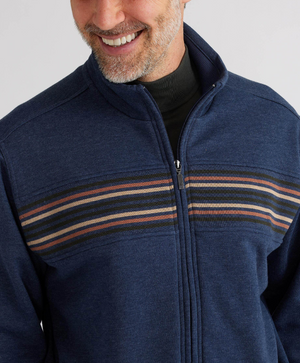 A person is wearing the Breakaway Bello Snowy Mt Fleece Jacket, a casual dark blue zip-up with horizontal multicolored chest stripes. Featuring a high collar, they smile slightly while only part of their face is visible.