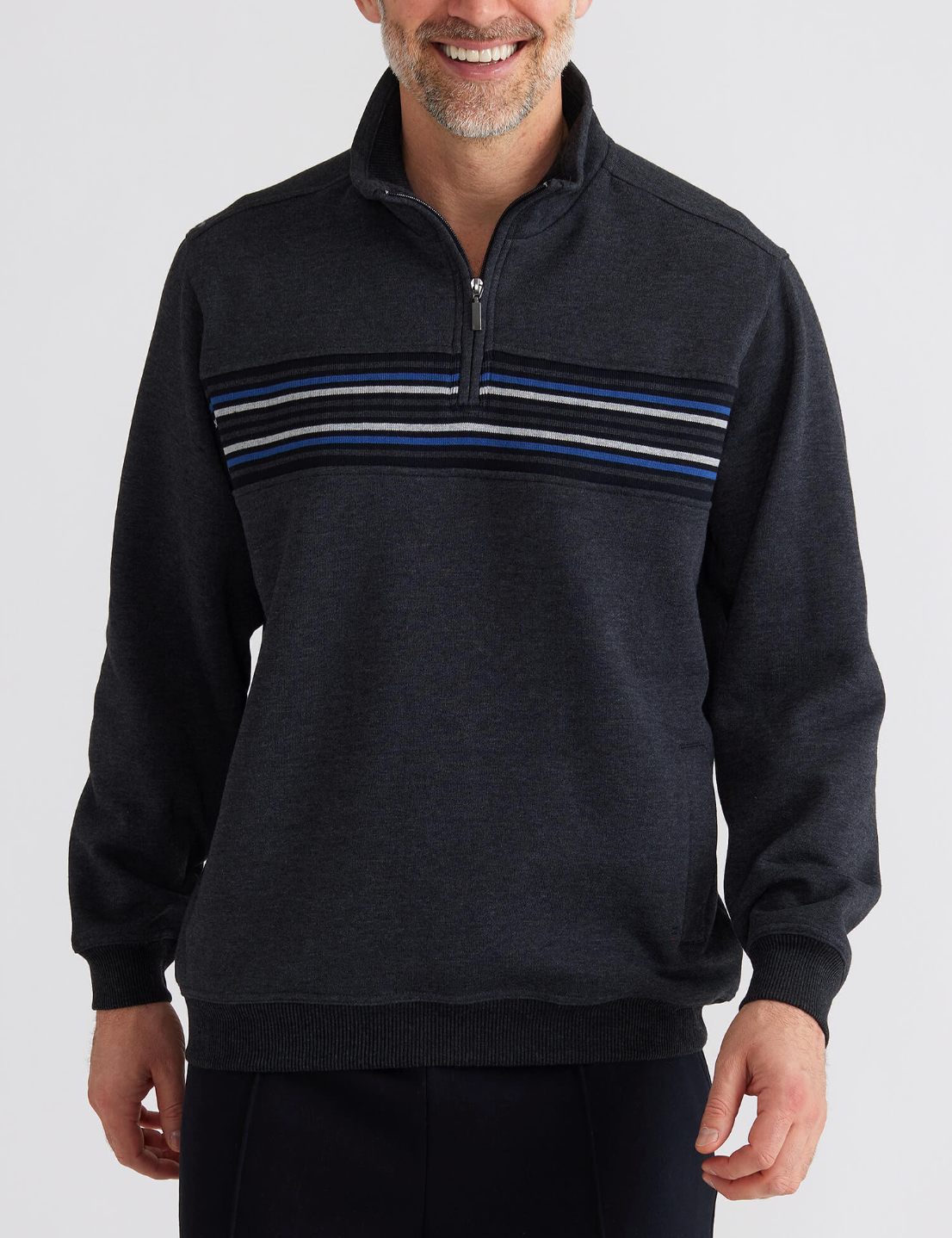 A man stands smiling against a white background, wearing the Breakaway Bello Snowy Mt Fleece Half Zip in dark gray with light gray, white, and blue stripes. The fleece offers comfort to his relaxed look as his hands rest by his sides.