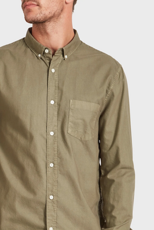 A man is showcasing an olive green Academy Brand Vintage Oxford Shirt with a chest pocket. His head is slightly turned to the left against a plain white background, drawing attention to the stylish shirt while only part of his face is visible.