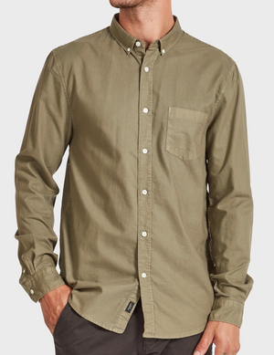 A person is wearing an olive green Academy Brand Vintage Oxford Shirt with a front pocket on the left side and long sleeves, paired with dark pants. The stylish shirt has white buttons and a collar. The person's left hand is in their pocket, and their face is not visible.