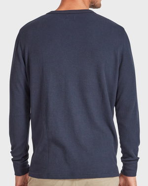 A person in the Academy Brand Workers Crew, a long-sleeve, navy blue shirt with rib knit texture and classic crew neck, paired with beige pants. The standard-fit shirt is viewed from behind as the person's hands rest calmly at their sides against a plain white background.