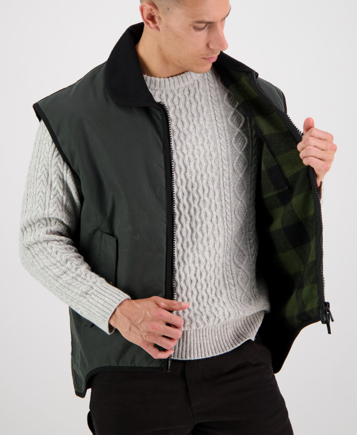 A person wearing a gray cable-knit sweater and a Swanndri Foxton Vest Wool Lined jacket with a black collar holds the jacket open to show its green plaid inner lining, looking down with a neutral expression.