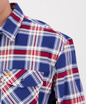 Close-up of a person wearing the Swanndri Egmont Flannelette shirt, featuring a blue, red, and white plaid pattern. Made of brushed cotton, this long sleeve shirt includes a buttoned chest pocket. The plain white background emphasizes its vibrant colors and design.