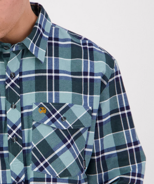 The image features a person wearing the Swanndri Egmont Flannelette, showcasing its blue and green plaid design with long sleeves, buttoned pockets, and collar in soft brushed cotton, capturing the cozy essence of this shirt.