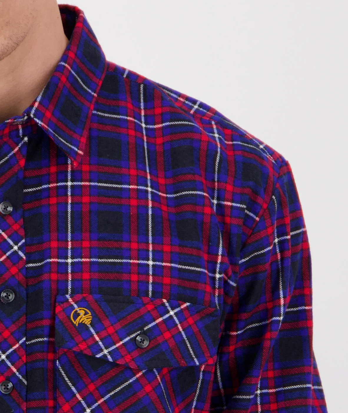 A person wears a Swanndri Egmont Flannelette shirt in blue and red plaid with a black button-up design. The long sleeve brushed cotton shirt includes a pocket adorned with an embroidered yellow logo. Only part of their face and upper torso is visible against a plain backdrop.