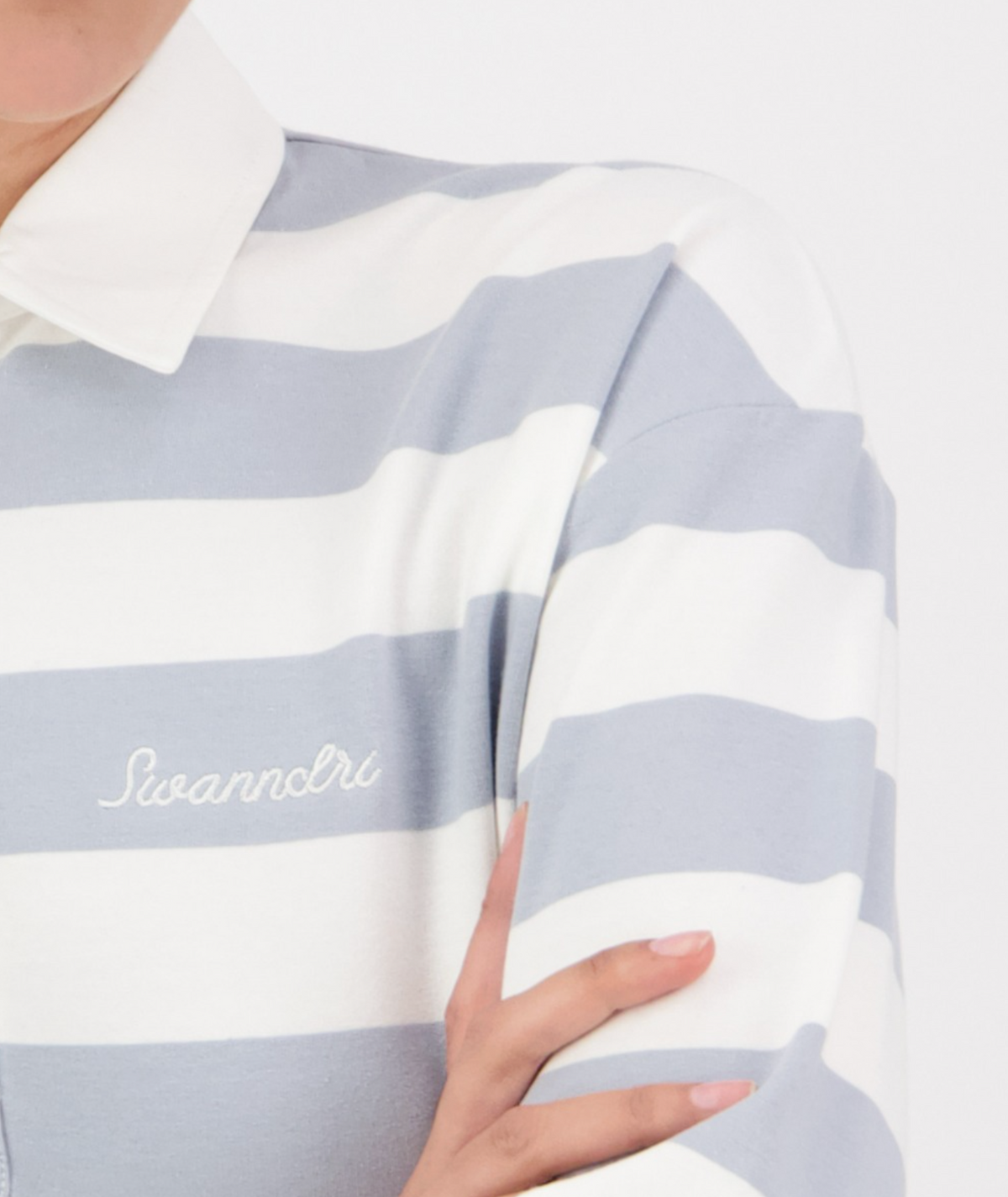 Wearing the Swanndri Carlaw Rugby shirt, a person exudes casual elegance in a crisp, white and light gray striped design with "Swanndri" embroidery. The stylish, sporty piece enhances their confident stance while only part of the torso and arms are visible.