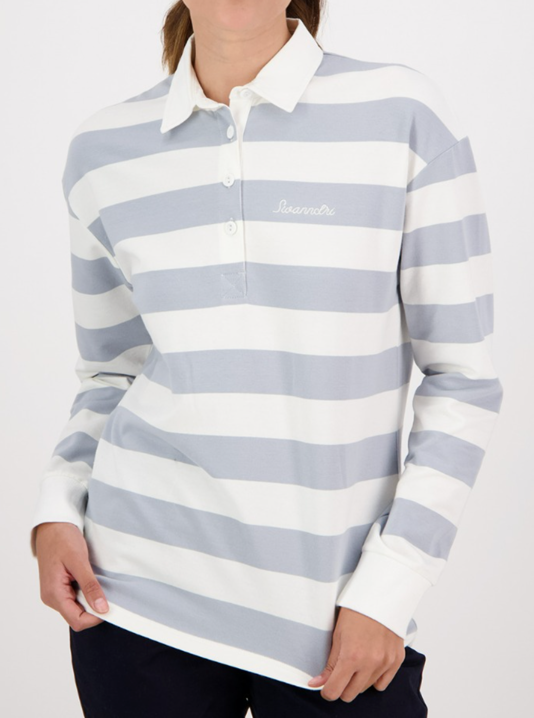 A person in a casual Swanndri Carlaw Rugby, a long-sleeve white and light gray striped polo with "Seanmoi" on the chest, evokes classic rugby style. They're paired with dark pants and have one hand in their pocket against a plain white background.