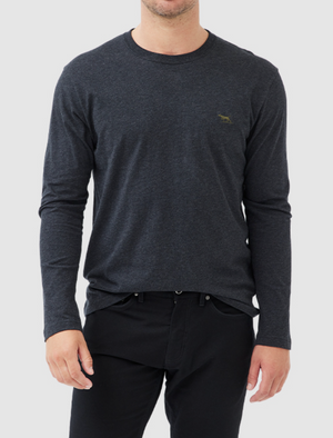 A person is wearing a Rodd & Gunn Long Sleeve Gunn T-Shirt, dark gray and 100% cotton, with a small embroidered logo on the left chest. The shirt has a sports fit and is paired with black pants against a light gray background. The person's face is not visible.