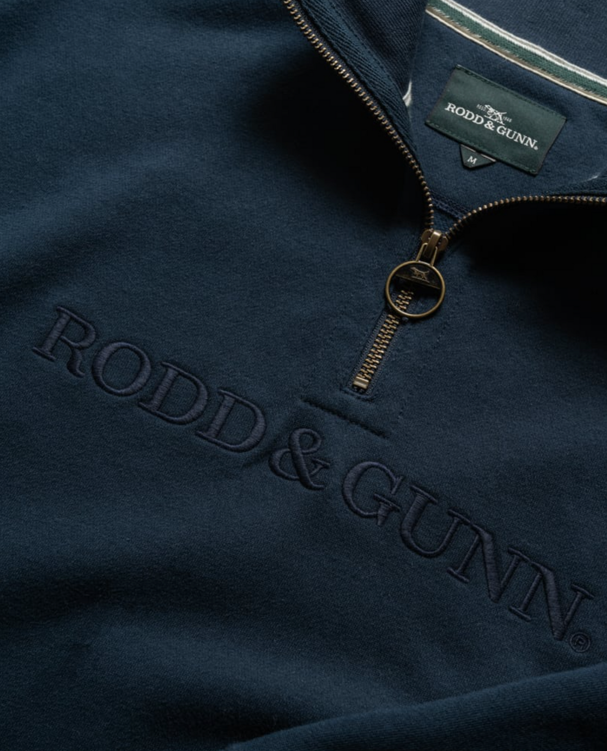 The close-up highlights the Rodd & Gunn Alford Forest Sweat, a navy mid-weight jersey pullover with a half zip. "RODD & GUNN" is subtly embroidered in matching thread, and the metal circular zipper pull reveals the tag with the brand's distinctive white and green logo.