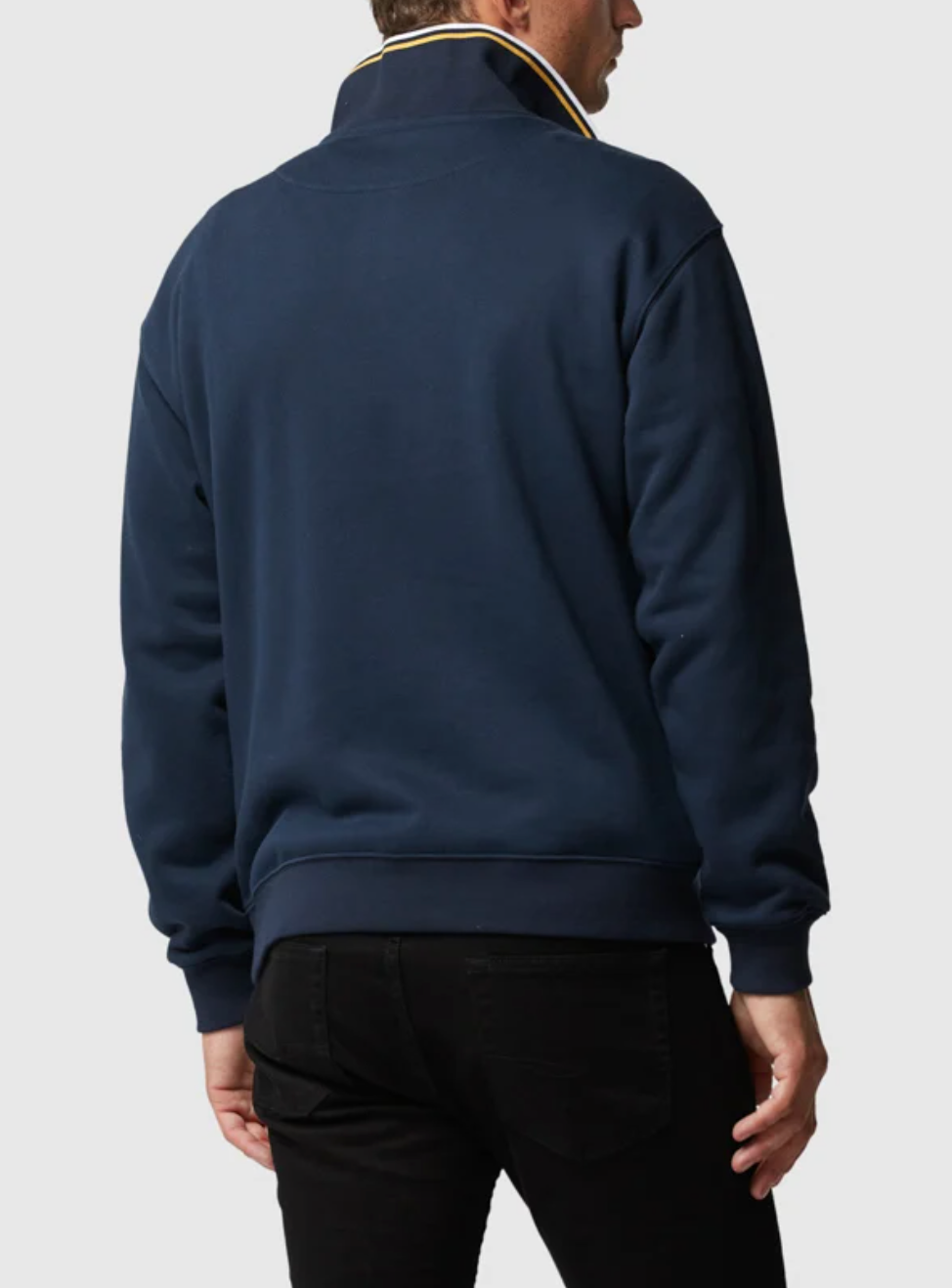 A man models the Rodd & Gunn Alford Forest Sweat, a navy blue jacket with mid-weight jersey, half-zip front, and high collar highlighted by yellow and white stripes. He pairs it with black pants, facing away against a plain light background.