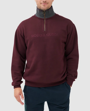 A man models the Rodd & Gunn Alford Forest Sweat, a burgundy half-zip sweater with the brand's embroidered logo, featuring a dark gray collar and zipper. Standing against a plain light gray background, he pairs it with dark pants for a casual yet sophisticated look.