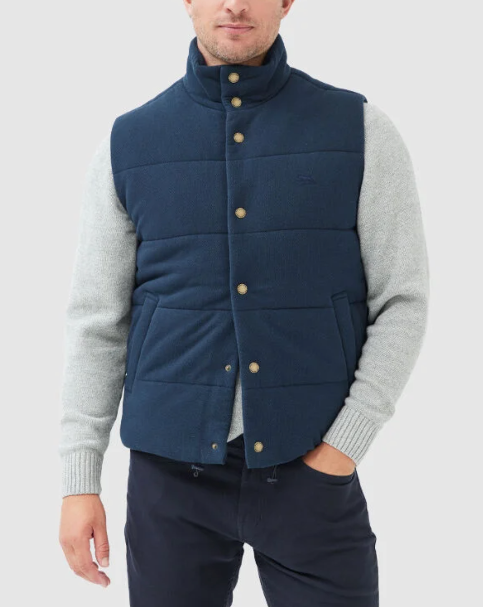 A man stands with his hand in his pocket, wearing the Rodd & Gunn Lake Ferry Vest—a navy blue padded vest with lightweight insulation, a high collar, and gold buttons—over a light gray sweater. The vest pairs perfectly with his dark trousers against the plain, light-colored background.