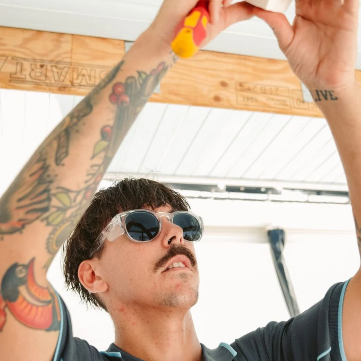 A person with a mustache and tattoos on their arms is wearing SafeStyle Cruisers Clear Frame Polarised Lens sunglasses along with safety goggles while using a tool overhead. The individual stands under a wooden structure and appears focused on their task.