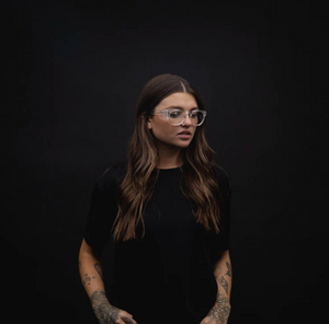 A woman with long brown hair, adorned with tattoos and wearing SafeStyle Cruisers Clear Frame Clear Lens glasses, stands against a dark background. Sporting a black shirt, she exudes both style and safety as she gazes to the side with a neutral expression.