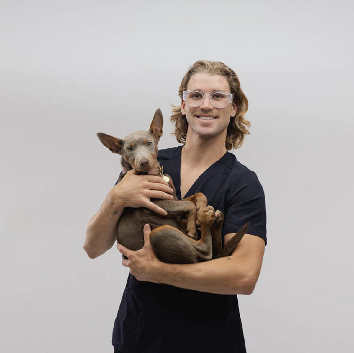 A long-haired person wearing SafeStyle Cruisers Clear Frame Clear Lens glasses smiles, holding a relaxed dog with pointy ears. They are in dark scrubs against a plain white background.