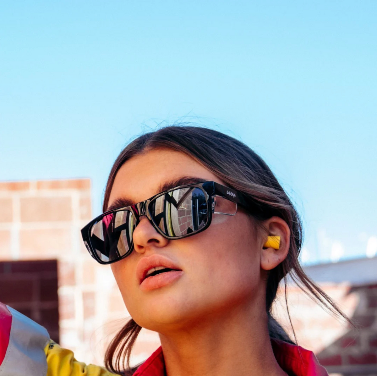 A woman, in SafeStyle Fusions sunglasses with UV-protected black frames, models outdoors against a blue sky. Yellow earplugs adorn her ears, while her long hair cascades over a colorful top, with a building backdrop enhancing the vivid scene.