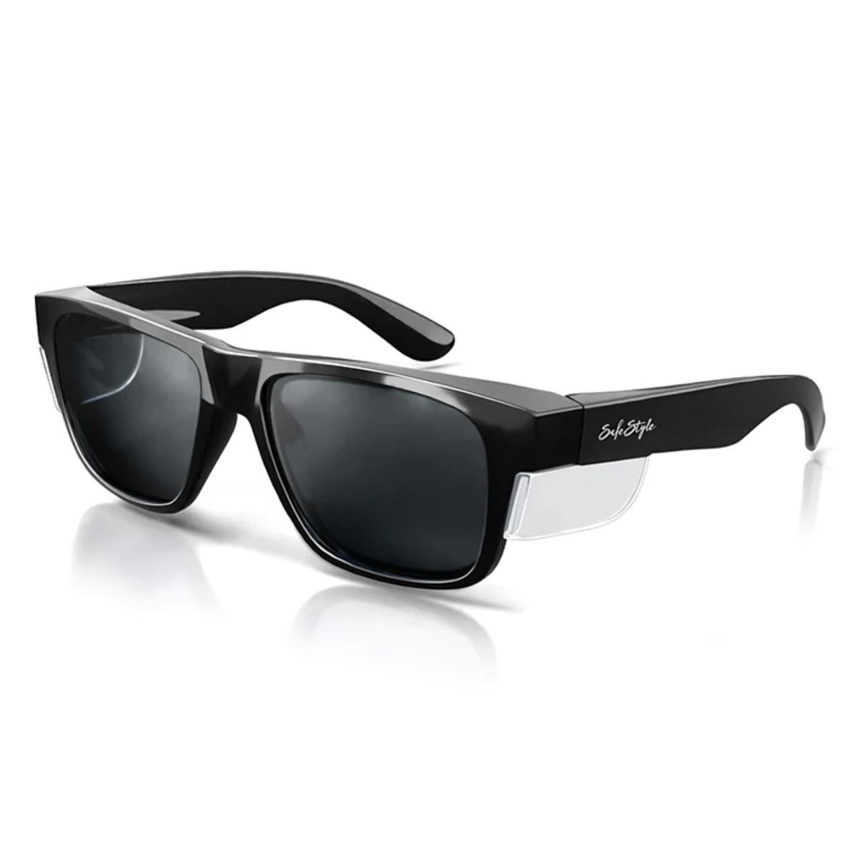 The SafeStyle Fusions Black Frame Polarised Lens sunglasses feature black side shields, dark lenses, and slightly curved arms. The frames have a glossy finish offering UV protection, with a small signature on the left temple. Resting on a reflective white surface, they blend style and safety.
