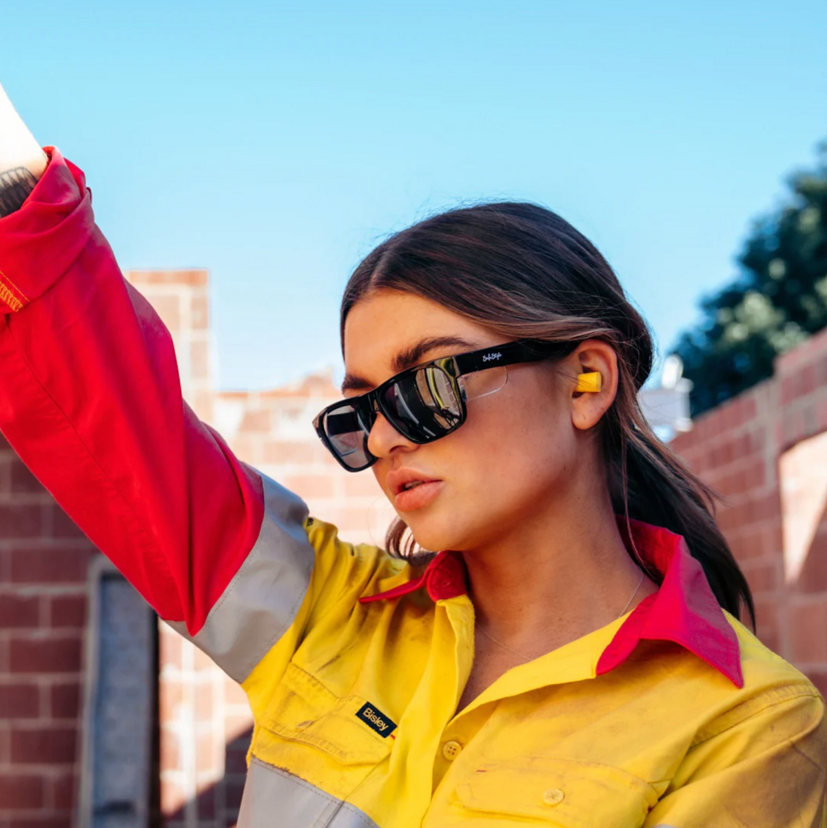 Wearing a high-visibility work shirt, a person stands confidently outside against a brick structure. With ear protection and raised arm under the clear blue sky, they wear SafeStyle Fusions Black Frame Tinted Lens sunglasses for added UV protection and safety.