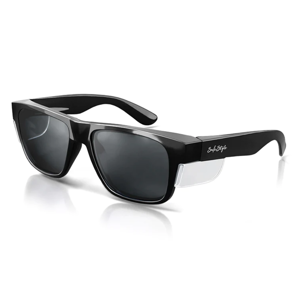 The SafeStyle Fusions Black Frame Tinted Lens sunglasses feature a graphite frame with dark lenses and clear side shields. A small temple logo subtly brands the style, enhanced by a reflective surface for a polished look, offering stylish UV protection against a white background.