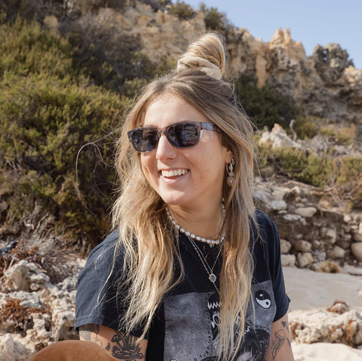 A woman with long, wavy blonde hair smiles outdoors in sunglasses. She wears a black T-shirt with a yin-yang design, layered necklaces, and hoop earrings. Her shades are the SafeStyle Classics Graphite Frame Tinted Lens, ideal for UV protection amid rocky terrain and greenery under clear skies.
