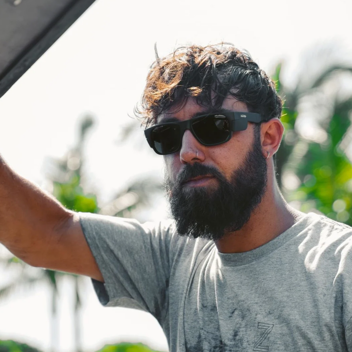 A bearded man wearing SafeStyle Classics Matte Black Frame Polarised Lens sunglasses with UV protection stands outdoors, holding something aloft against a backdrop of palm trees and a clear sky.
