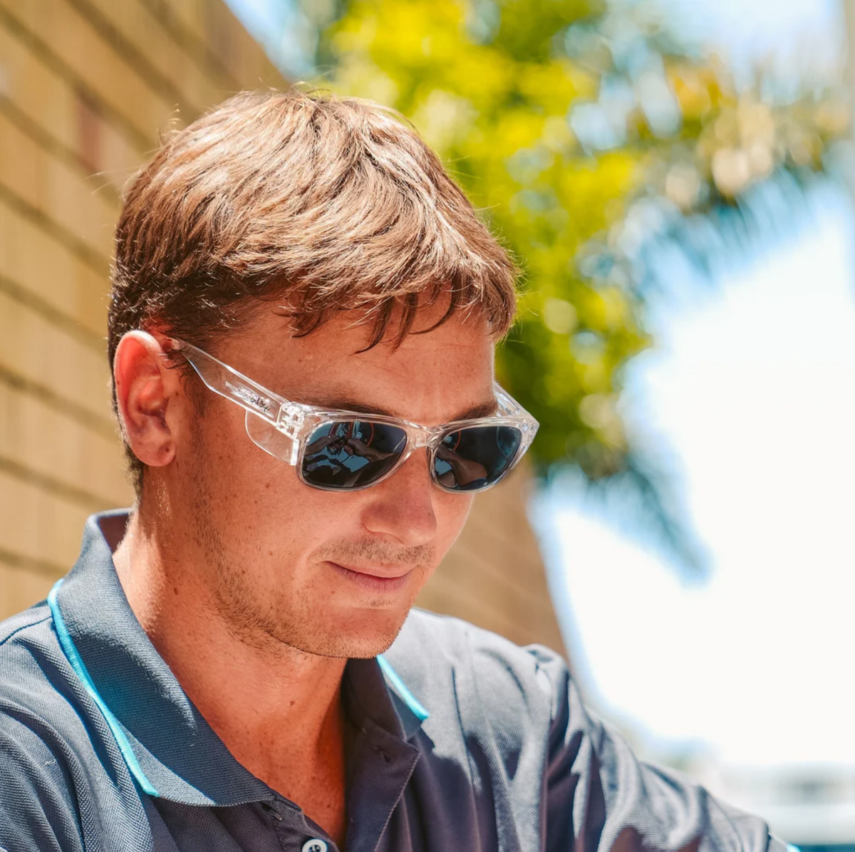 A man stands outdoors wearing SafeStyle Classics Clear Frame Polarised Lens sunglasses and a gray collared shirt. With short brown hair, he is near a brick wall, and the blurred background features a green-leaved tree and a clear blue sky.