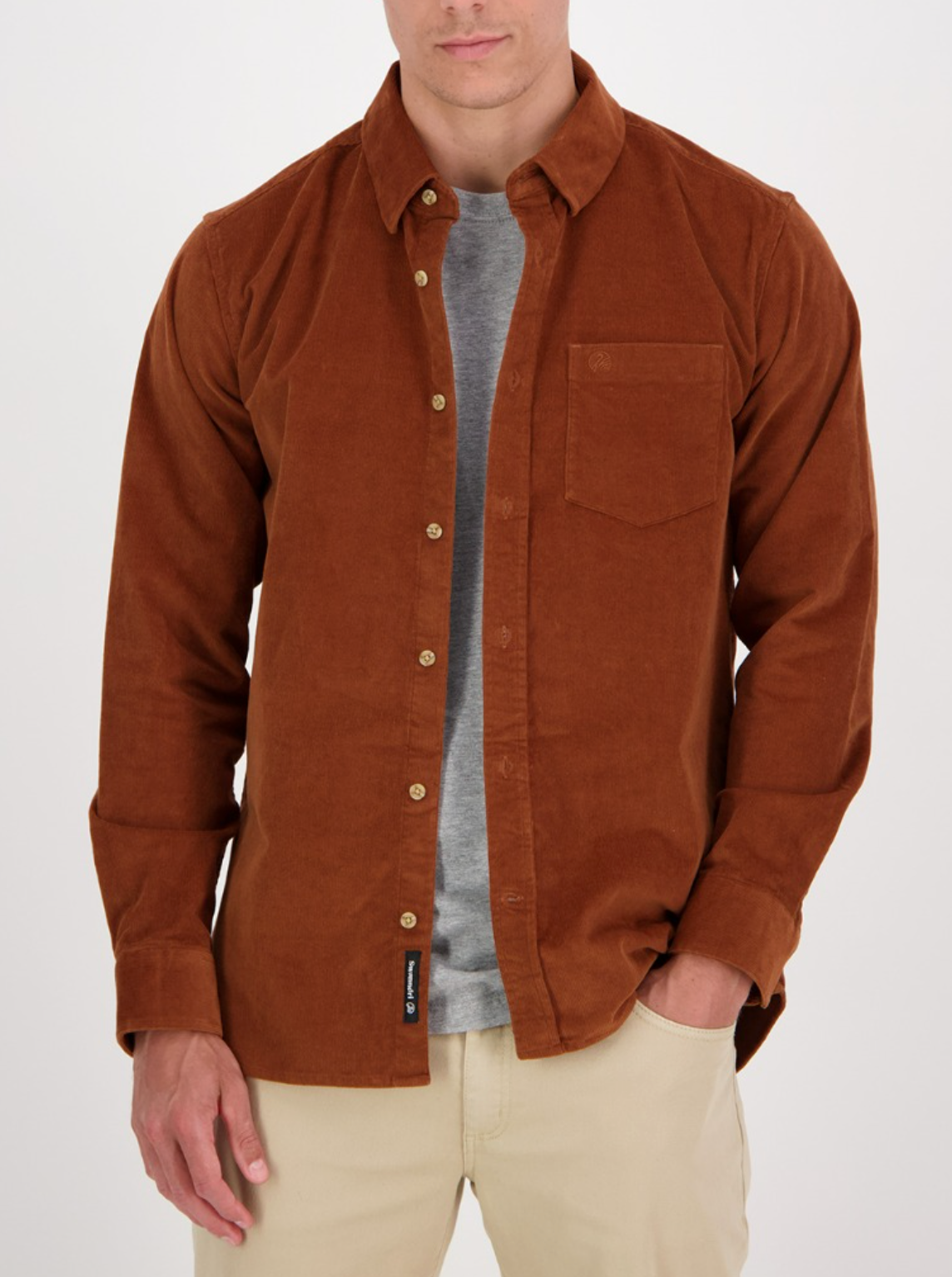 Someone is wearing a Swanndri Ranfurly Corduroy Shirt in brown over a gray t-shirt with beige pants, one hand in their pocket against a plain backdrop. The shirt's visible chest pocket adds to its stylish look.