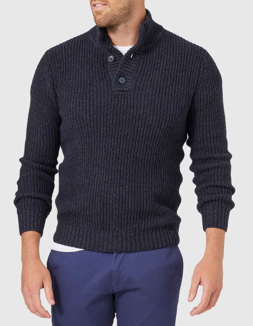 Wearing the Blazer Oscar Button Neck Knit, a dark ribbed sweater with ribbed cuffs and waist, paired with dark blue pants, the person stands out against the plain light background. Their head is slightly turned, concealing their features.