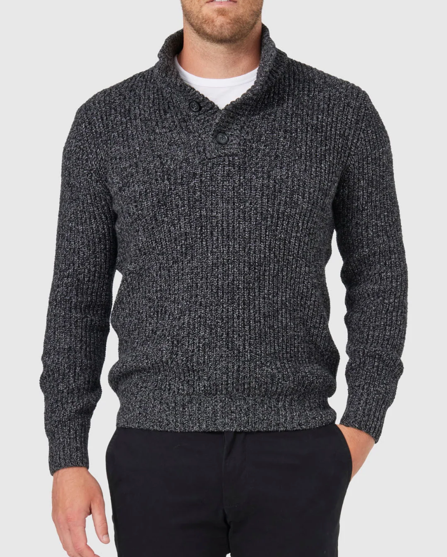 Wearing a Blazer Oscar Button Neck Knit in dark gray, a person stands against a light gray background with one hand in the pocket of their dark pants.