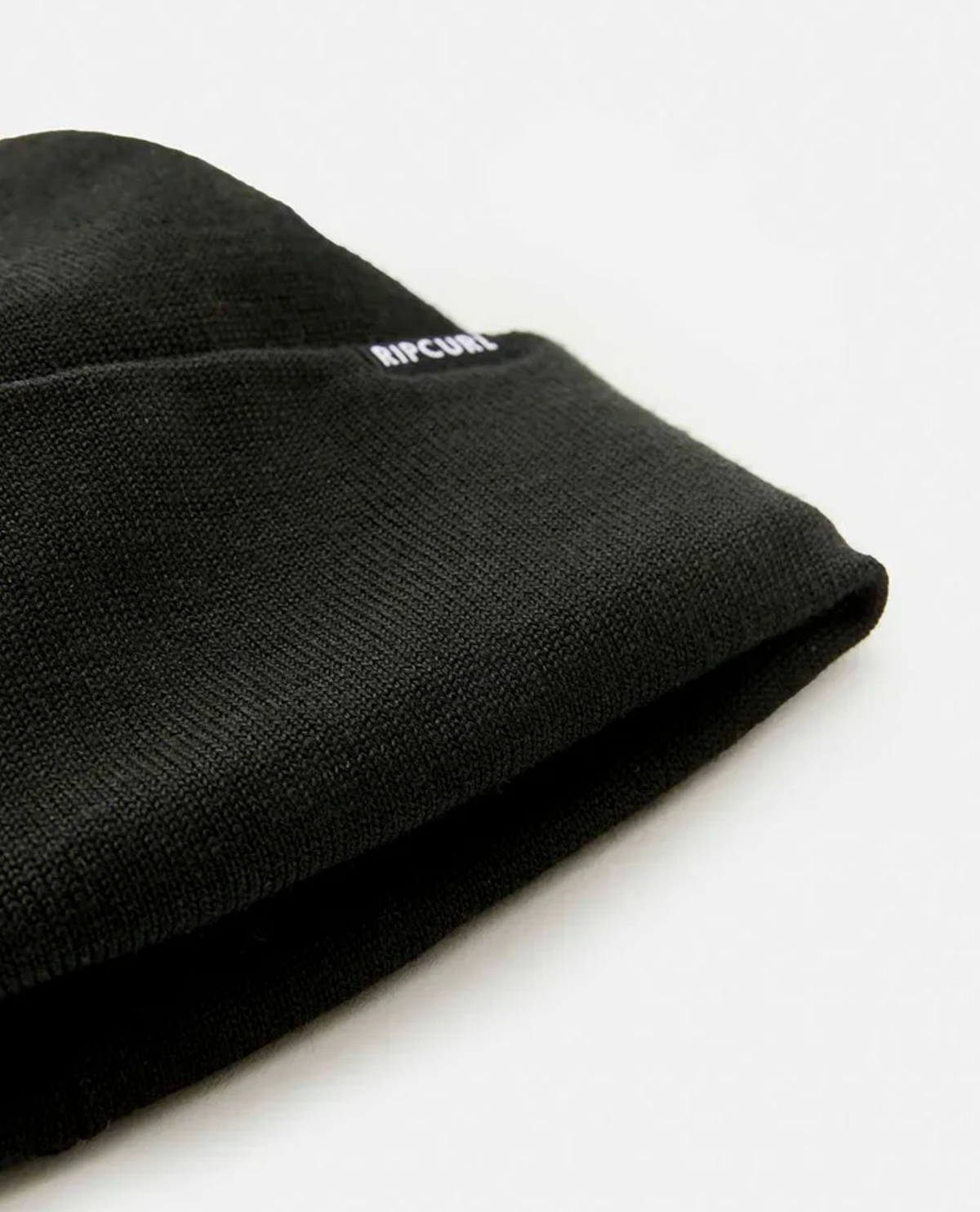 A close-up of the Rip Curl Anti-Series Regular Beanie against a white background. The textured black knit fabric adds depth, with "RIP CURL" subtly printed in small white letters near the edge, promising style and warmth.
