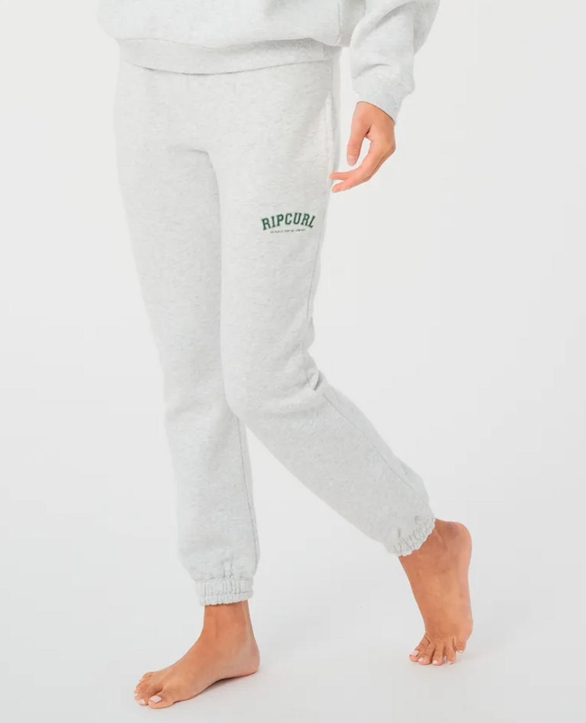 A person wearing Rip Curl Varsity Pants, featuring "Rip Curl" in green on the left leg, stands barefoot on one foot with the other slightly bent. The light gray trackpants offer comfort with a fleece lining against the plain white background.