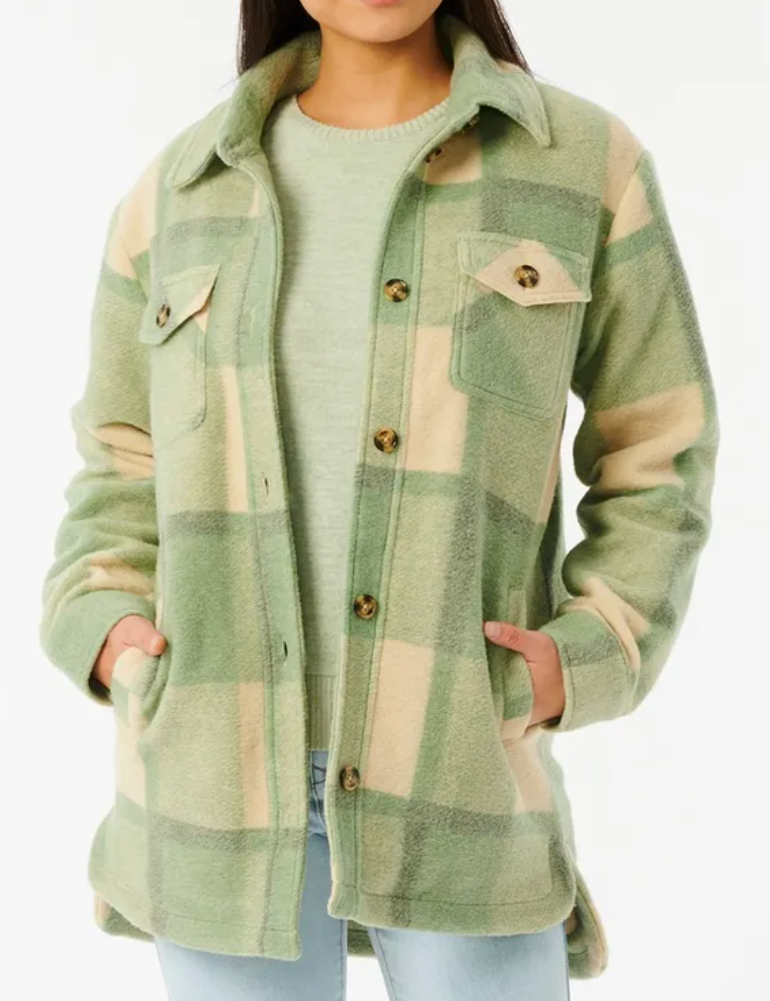 Someone is wearing the Rip Curl Maniciana Jacket, a stylish green and beige plaid fleece with front button closure and chest pockets. Its quilted lining adds warmth. Layered over a light green top, they have their hands comfortably in the pockets.