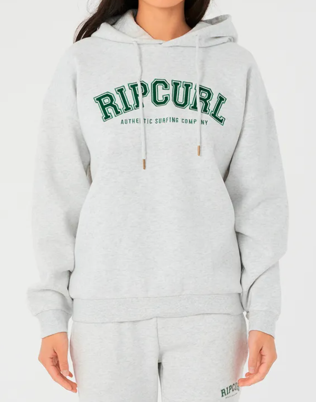 A person in a light gray Rip Curl Varsity Hoodie with relaxed fit and matching sweatpants. The hoodie showcases bold green "RIP CURL" lettering and has a cozy fleece lining, all set against a plain white backdrop, highlighting the comfort and style of this casual outfit.