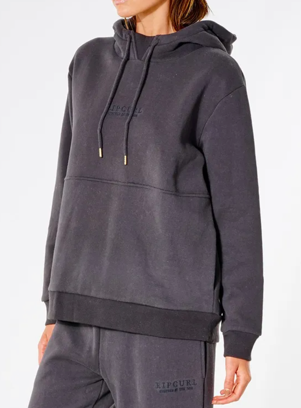 Someone wears a comfortable, dark gray Rip Curl Premium Surf Hoodie with matching sweatpants. The relaxed-fit hoodie and pants feature subtle text on the chest and near the pocket, set against a plain white background.