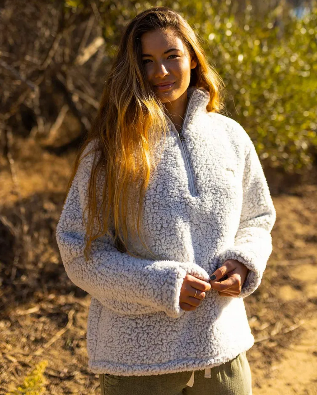 A person with long hair stands outside, wearing a Rip Curl Dark N Stormy 1/4 Zip Crew fleece jumper. Sunlit foliage and dry grasses form the background as they look towards the camera with a slight smile.