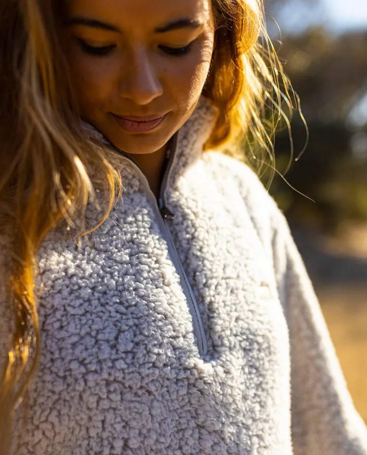 A person with long hair looks down, wearing the cozy Rip Curl Dark N Stormy 1/4 Zip Crew made from super soft fleece. The background is softly blurred with warm, natural light, perfect for this Rip Curl-inspired look.
