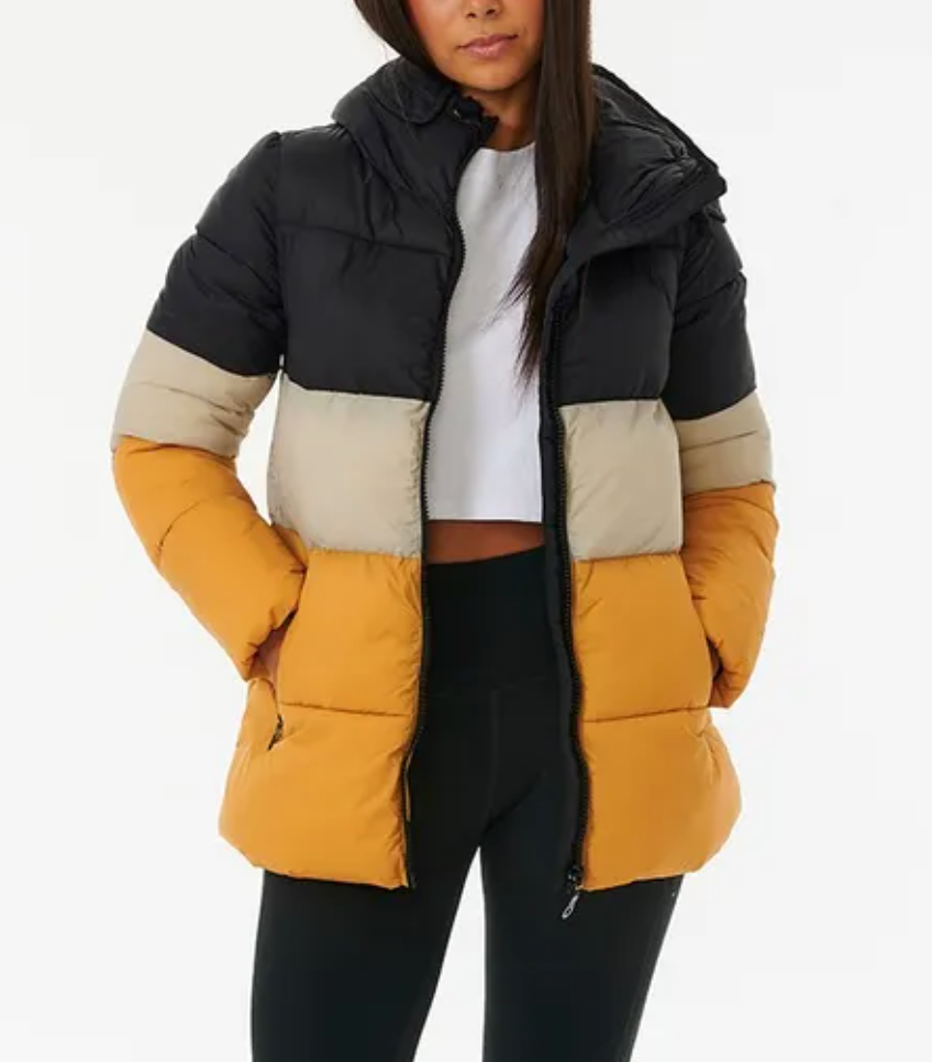 A person wears a Rip Curl Anti Series Insulated II Jacket with black, beige, and mustard stripes. This DWR water-repellent puffer is half-zipped, complementing their long hair, white cropped top, and black leggings in a neutral setting.
